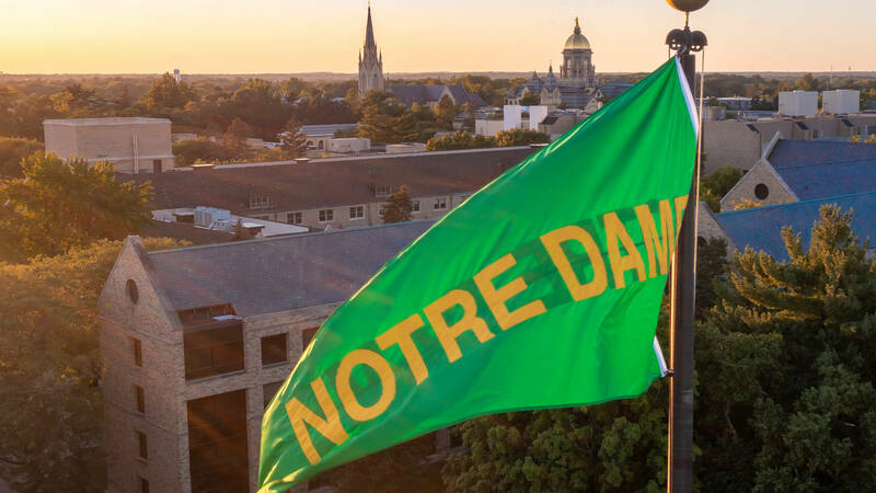 A green Notre Dame flag curls in the breeze atop the stadium. Lit by the light of a setting sun. The campus can be seen behind the flag.